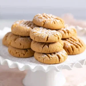A stack of soft gluten free peanut butter cookies with a classic criss-cross pattern, lightly dusted with powdered sugar and served on a white cake stand.