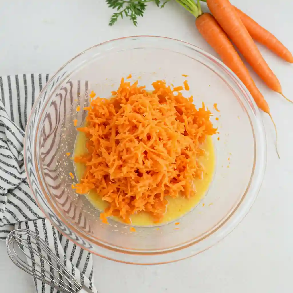 The process of making gluten-free carrot cake: Finely shredded carrots are added to the liquid ingredients in a glass mixing bowl, with a whisk and whole carrots visible in the background on a white countertop