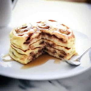 A golden-brown stack of Cinnamon Roll Pancakes topped with a generous swirl of thick cream cheese glaze, ready to serve for breakfast.