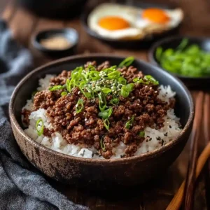 A rustic bowl featuring one of the easiest Korean Ground Beef Recipes, showing caramelized beef bulgogi on rice with chopsticks and a fried egg in the background.