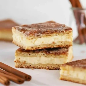 Stack of three golden brown Churro Cheesecake bars coated in cinnamon sugar on a white plate.